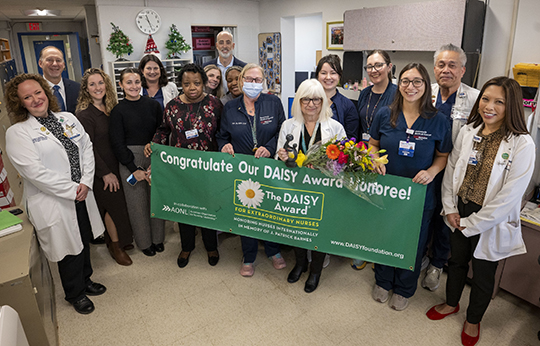 Carole is shown holding her DAISY Award trophy and the signed DAISY Award banner