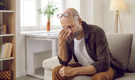 man sitting on couch holding his hand to his face, dealing with fatigue