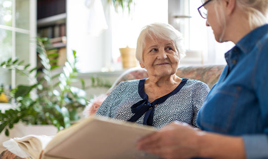 Senior woman and her adult daughter looking at photo album together on couch in living room