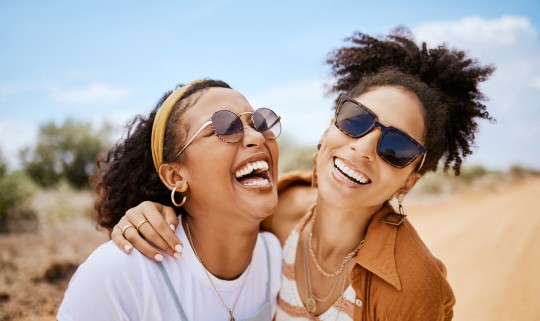 two women standing outside, wearing sunglasses, hugging and laughing
